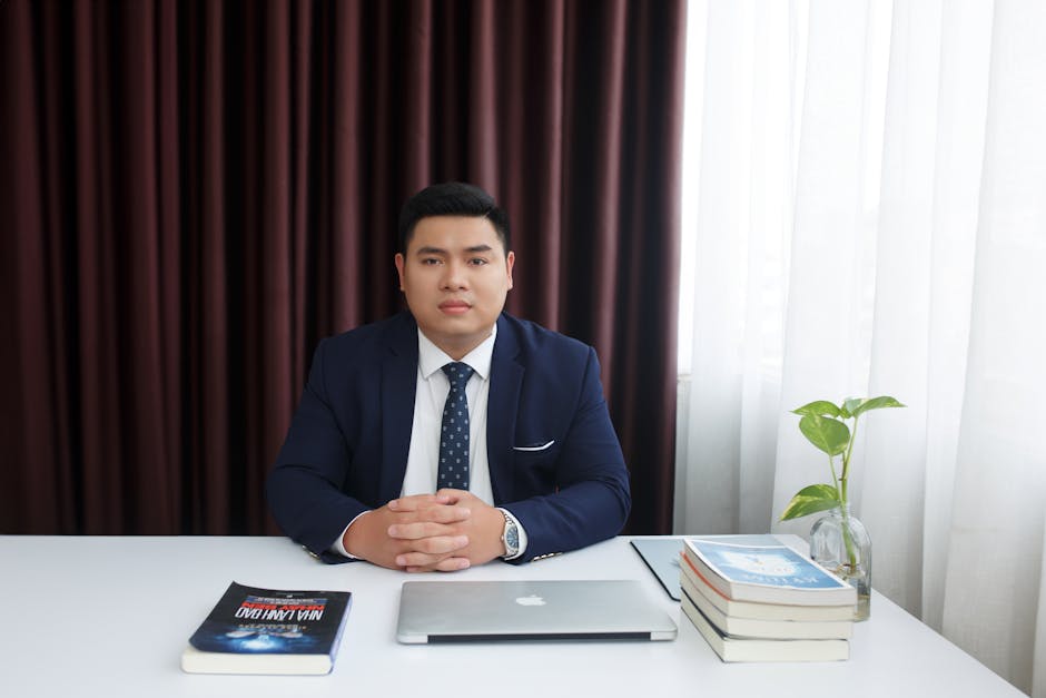 Businessman in a formal suit sitting at a desk with a laptop and books in a modern office setting.