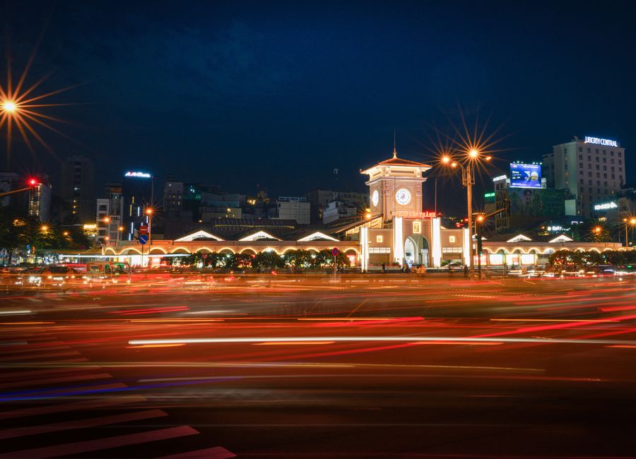 A dynamic long-exposure shot of Ben Thanh Market's illuminated facade in Ho Chi Minh City at night.