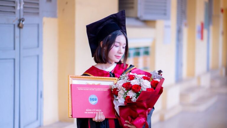 A young woman in a graduation cap and gown holds a certificate and bouquet, symbolizing academic achievement.