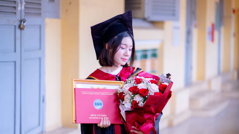 A young woman in a graduation cap and gown holds a certificate and bouquet, symbolizing academic achievement.