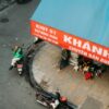 A person on a motorcycle beside people sitting on stools under a red awning