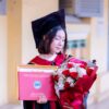 A young woman in a graduation cap and gown holds a certificate and bouquet, symbolizing academic achievement.
